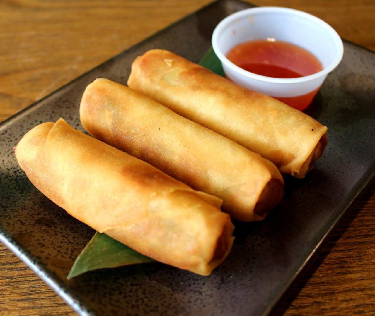 Top-down view of three fried spring rolls with a side of red sauce on a dark ceramic plate.