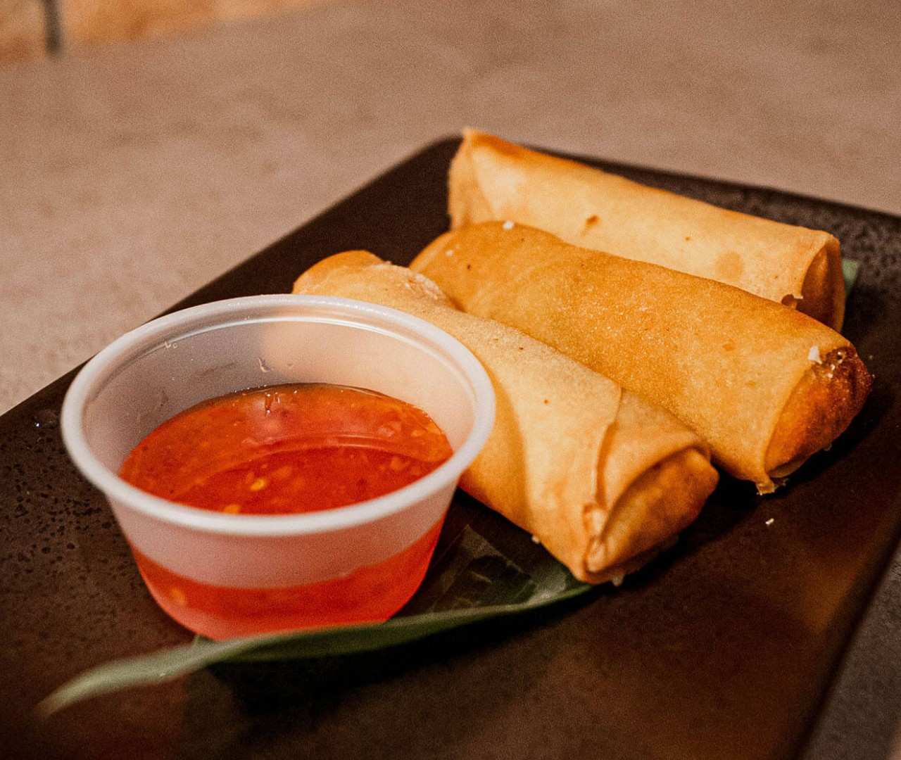 Three golden fried spring rolls arranged on a black rectangular plate with sweet chili dipping sauce.