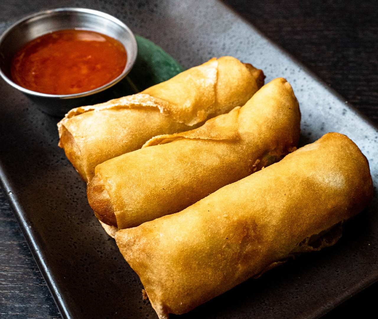 Three appetizer rolls on a dark textured plate with chili sauce, typical of Asian cuisine in Miami.