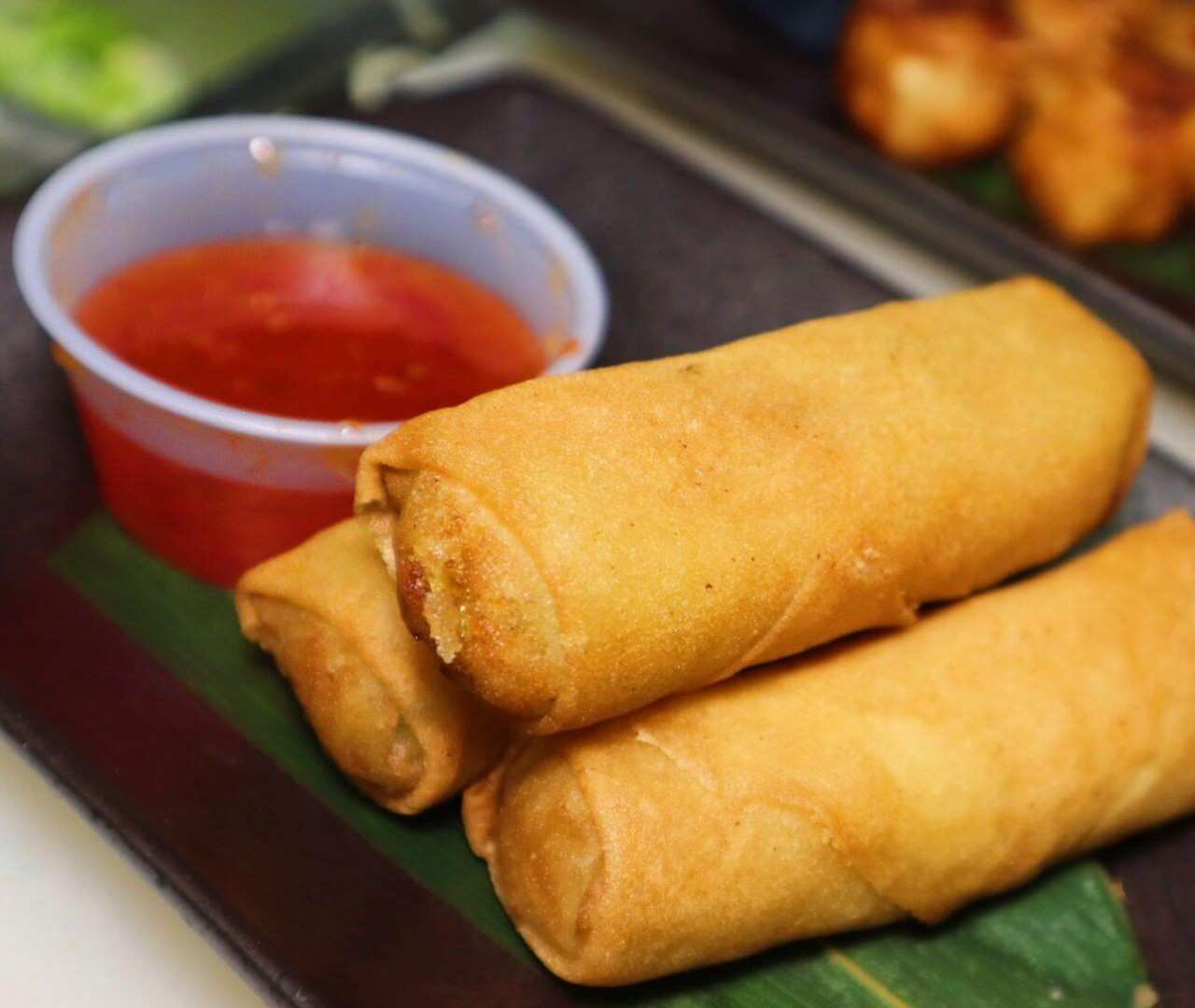Stack of crispy vegetable spring rolls served on a banana leaf with red dipping sauce.